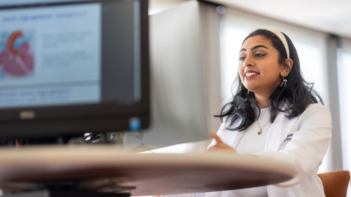 A medical student wearing a white coat types on a computer in a study room