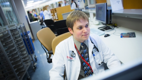 A resident wearing a white coat works at a computer in the hospital