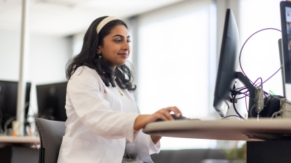 A medical student wearing a white coat types on a computer in a study room