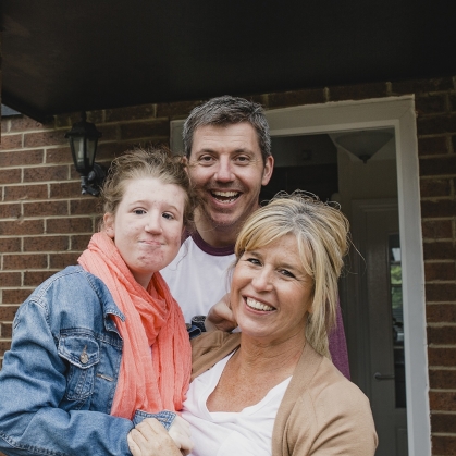 A family of three stands on a porch of their brick home