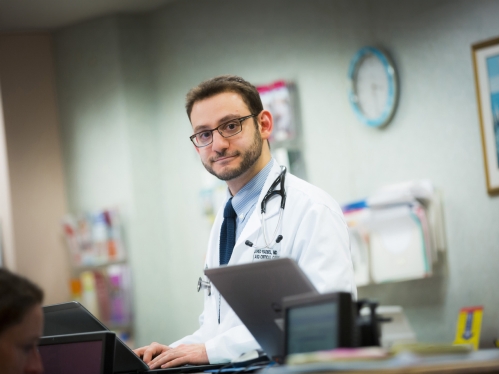 A resident wearing a white coat works at a computer in the hospital